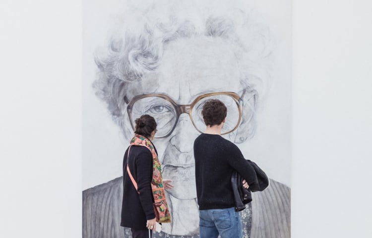 Two people viewing a painting of an older woman in glasses.