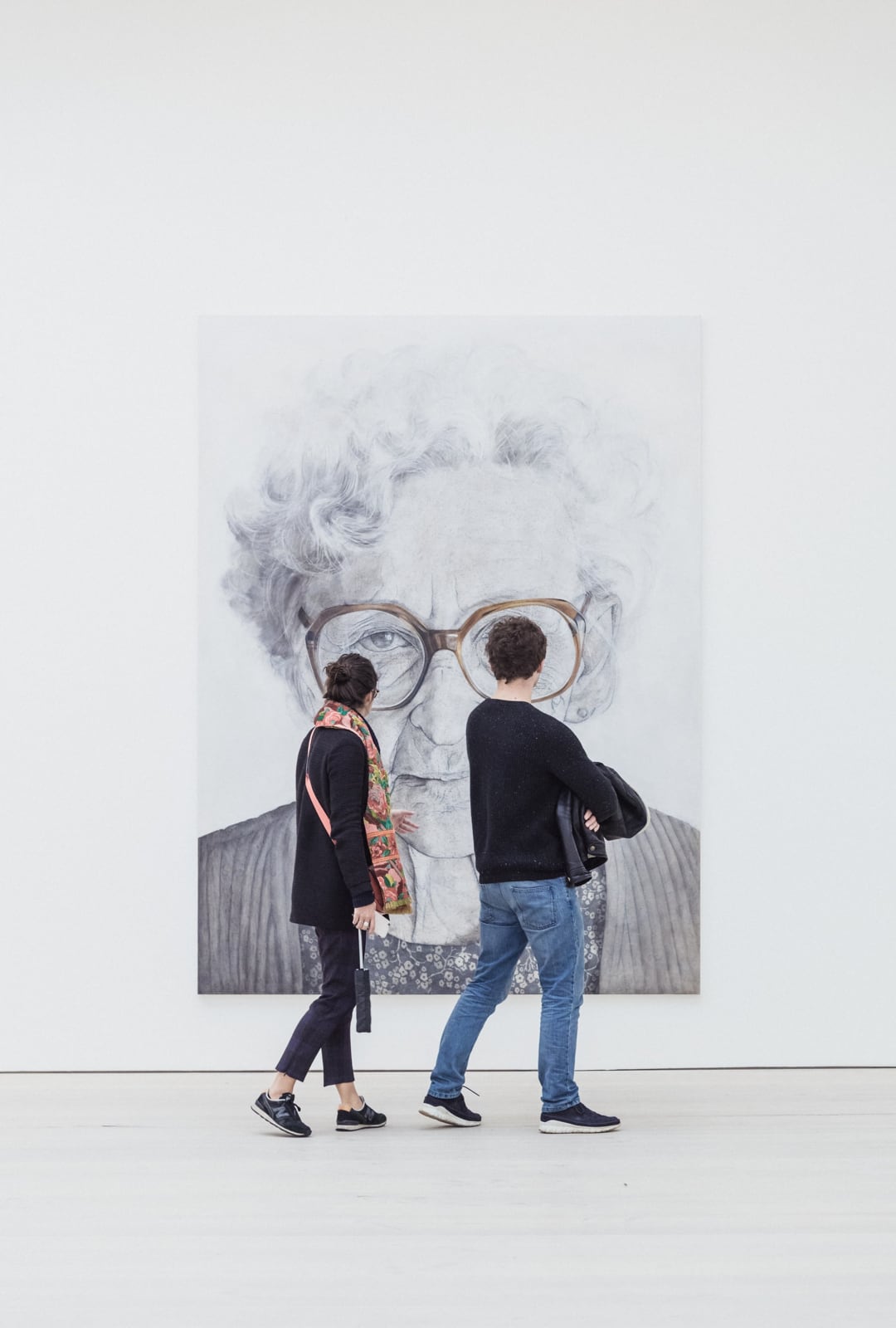 Two people viewing a painting of an older woman in glasses.