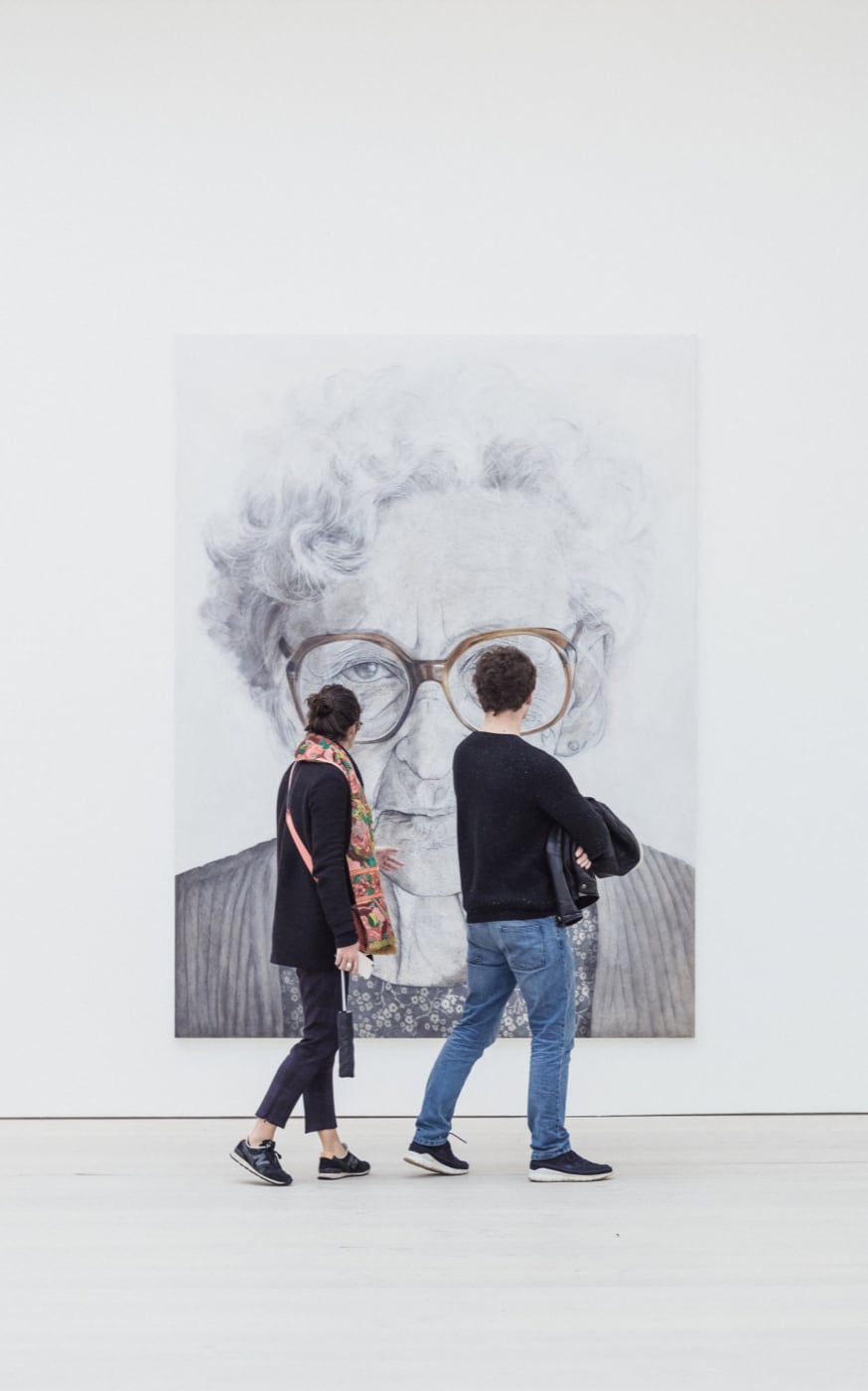 Two people viewing a painting of an older woman in glasses.